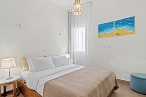Un dormitorio con una cama, una mesita de noche con lámpara, un reposapiés azul y dos cuadros con motivos marinos en la pared.