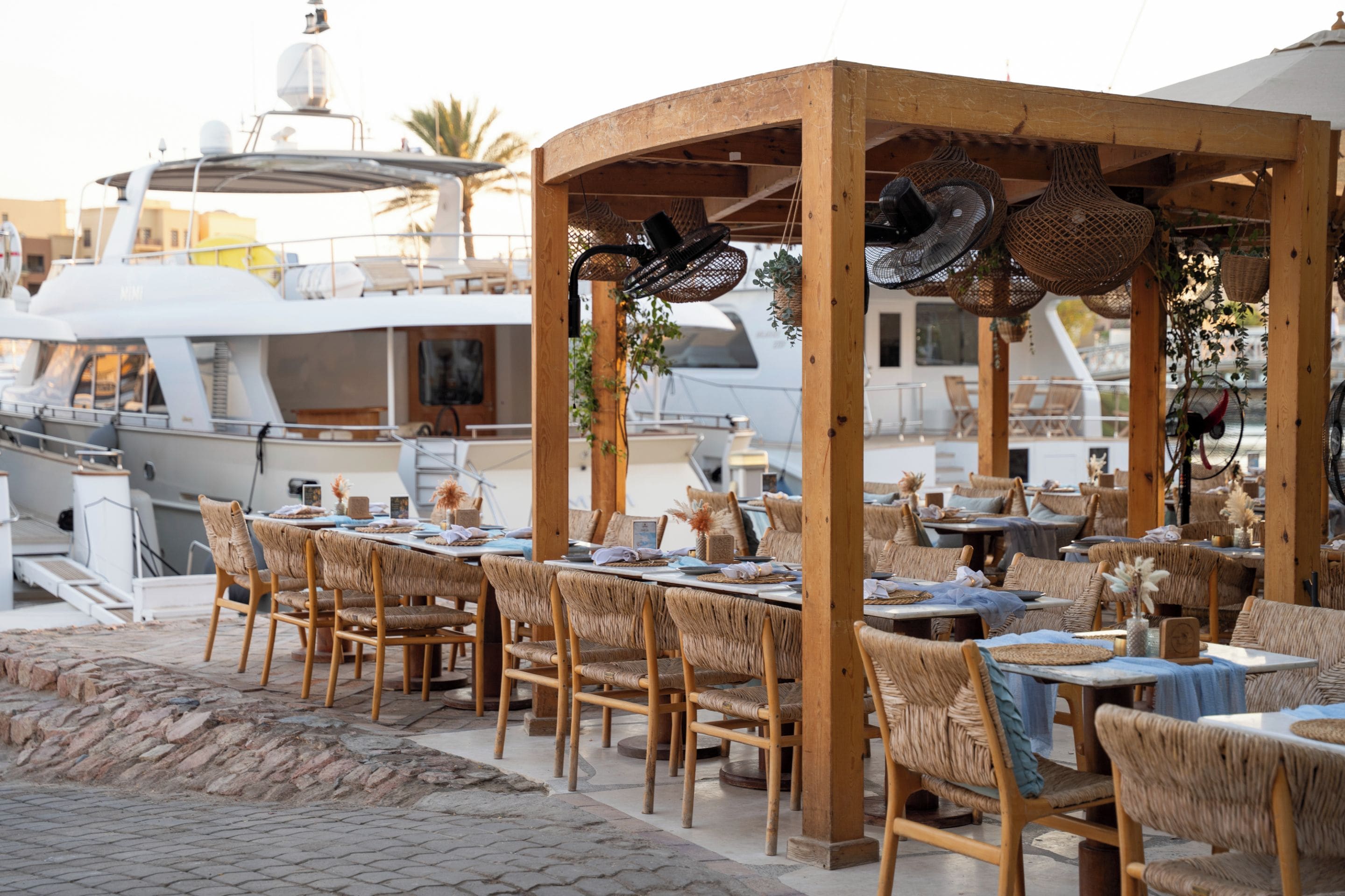An outdoor dining area with wicker furniture and hanging lights, overlooking a marina with yachts.