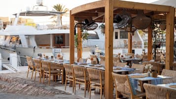 An outdoor dining area with wicker furniture and hanging lights, overlooking a marina with yachts.