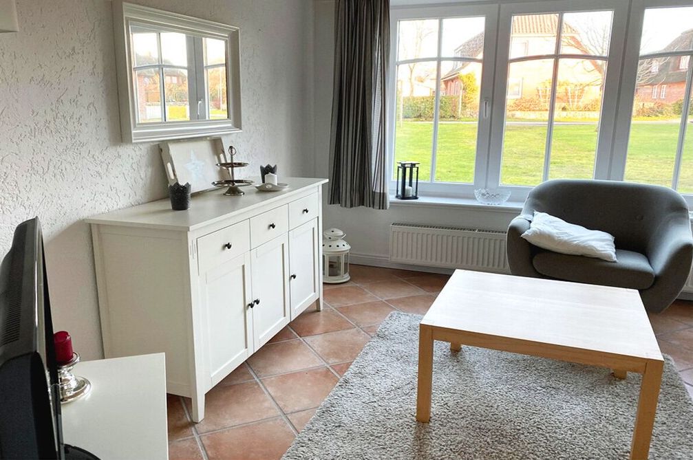 A room with a white cabinet, a grey armchair, a wooden table, and a view of a green lawn through large windows.