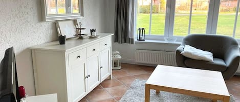 Una habitación con un armario blanco, un sillón gris, una mesa de madera y vistas a un césped verde a través de amplias ventanas.