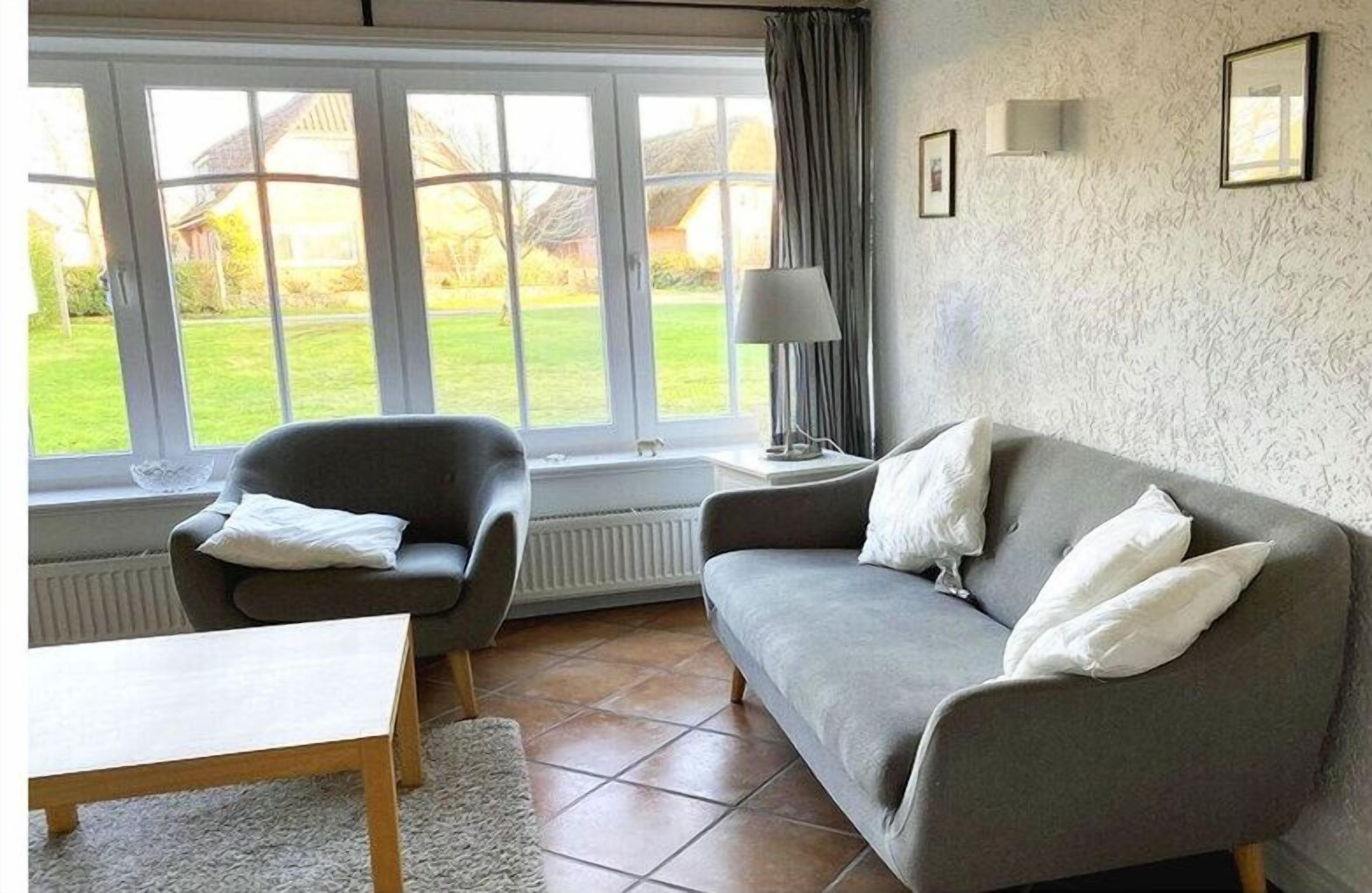 A living room with a sofa, armchair, and a table. There is a window with a view of a garden.