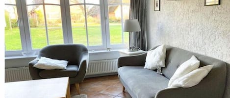 A living room with a sofa, armchair, and a table. There is a window with a view of a garden.
