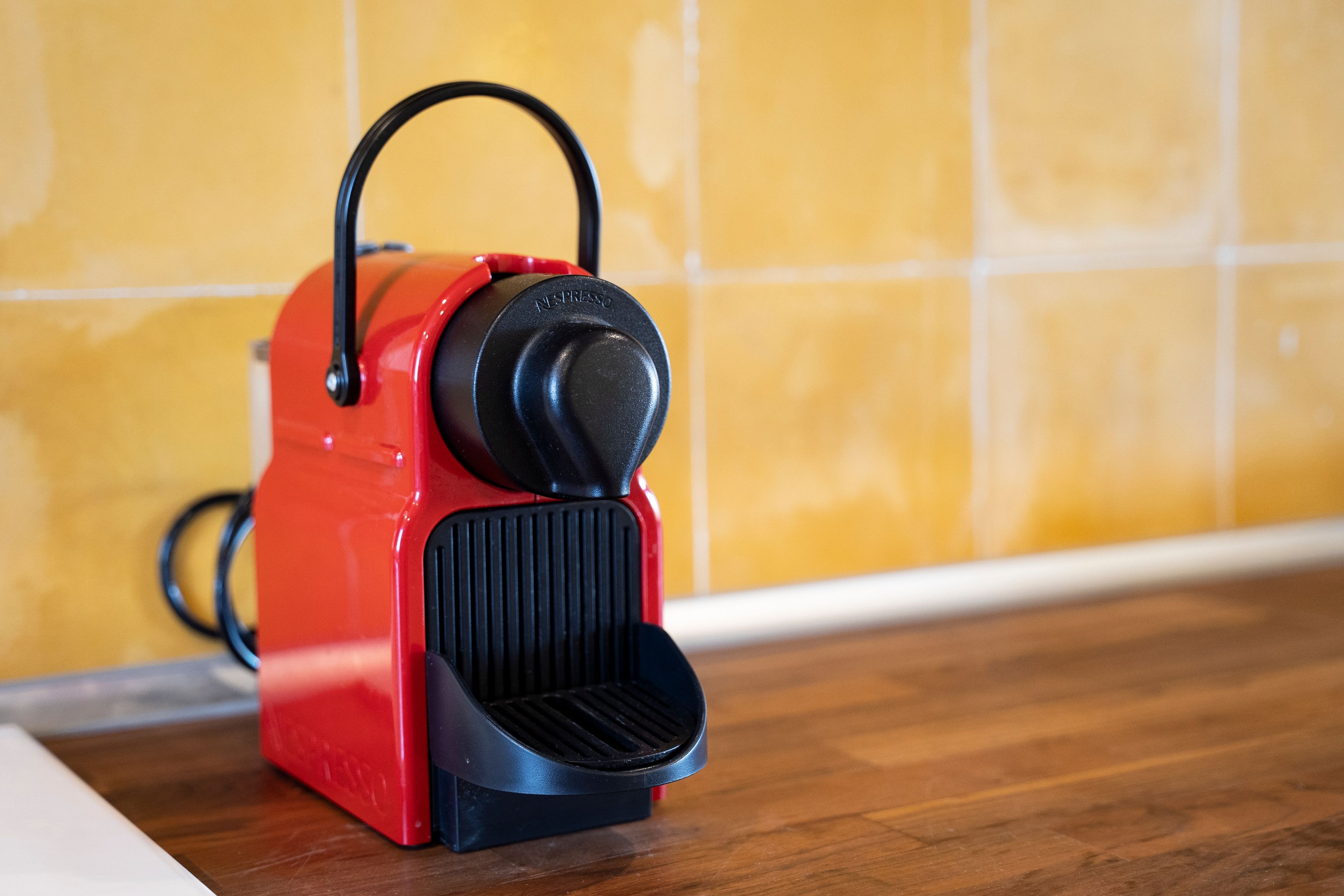 A red portable espresso machine with a black handle and a black base, placed on a wooden surface against a yellow tiled wall.