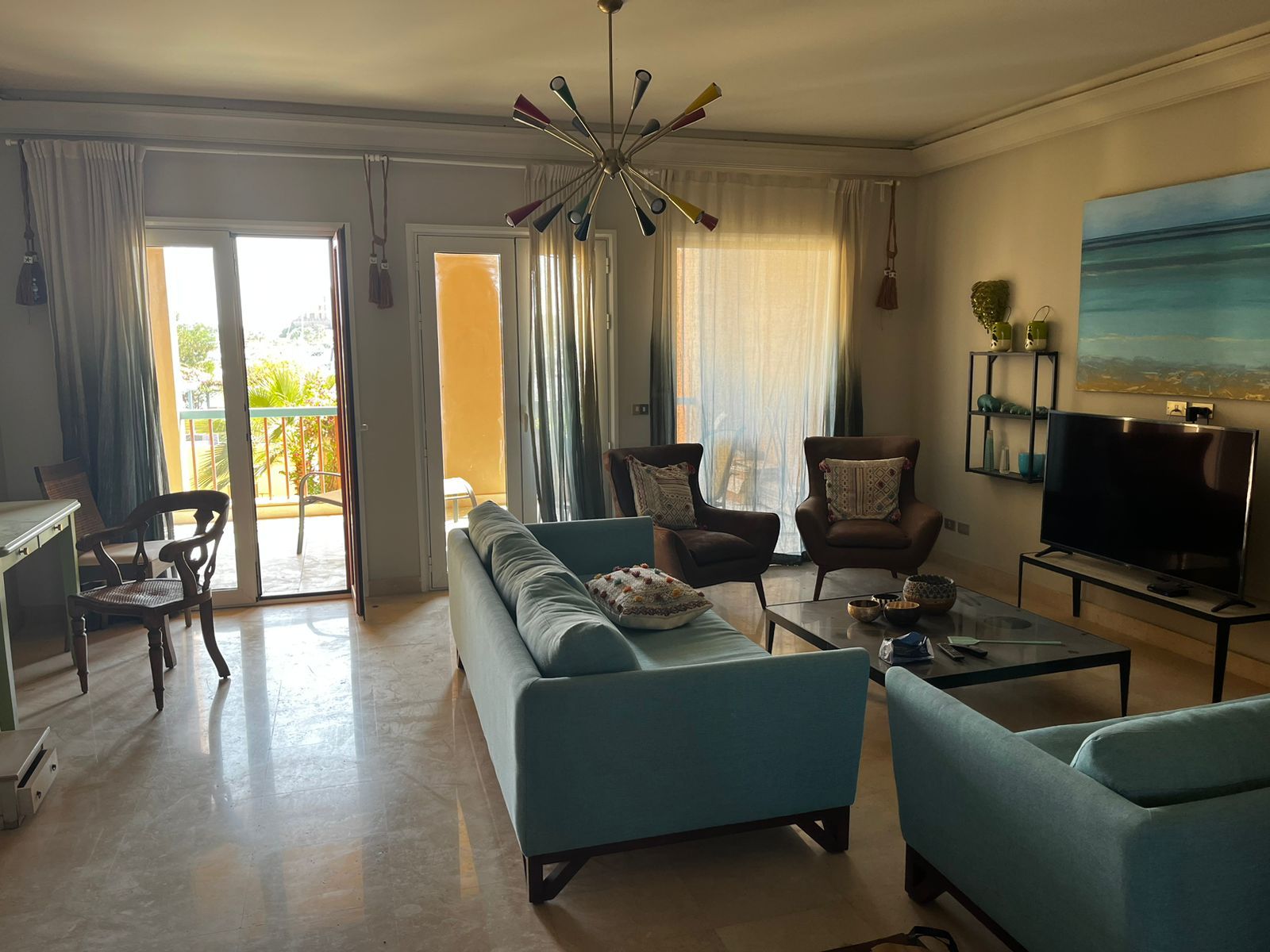 A living room with a sofa, armchairs, a coffee table, a TV, and a balcony with curtains.
