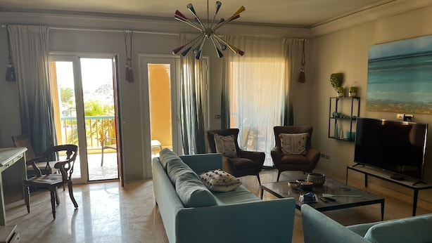 Sala de estar com sofá, poltronas, mesa de centro, televisão e varanda com cortinas.