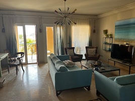 Una sala de estar con sofá, sillones, una mesa de centro, televisor y un balcón con cortinas.