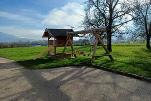 Ein hölzernes Schaukelgerüst mit einer kleinen Schutzhütte, einem gepflasterten Weg und einer Rasenfläche mit Bäumen und Gebäuden in der Ferne.