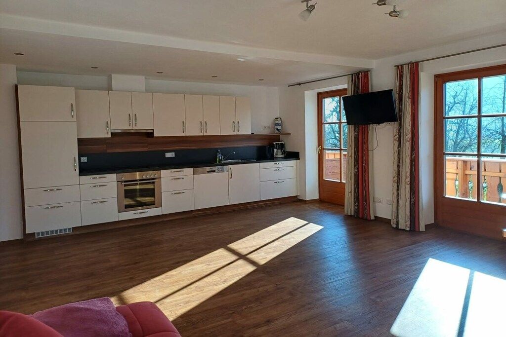 A modern kitchen with white cabinets, a black countertop, and a built-in oven. There is a large window with curtains and a television mounted on the wall.