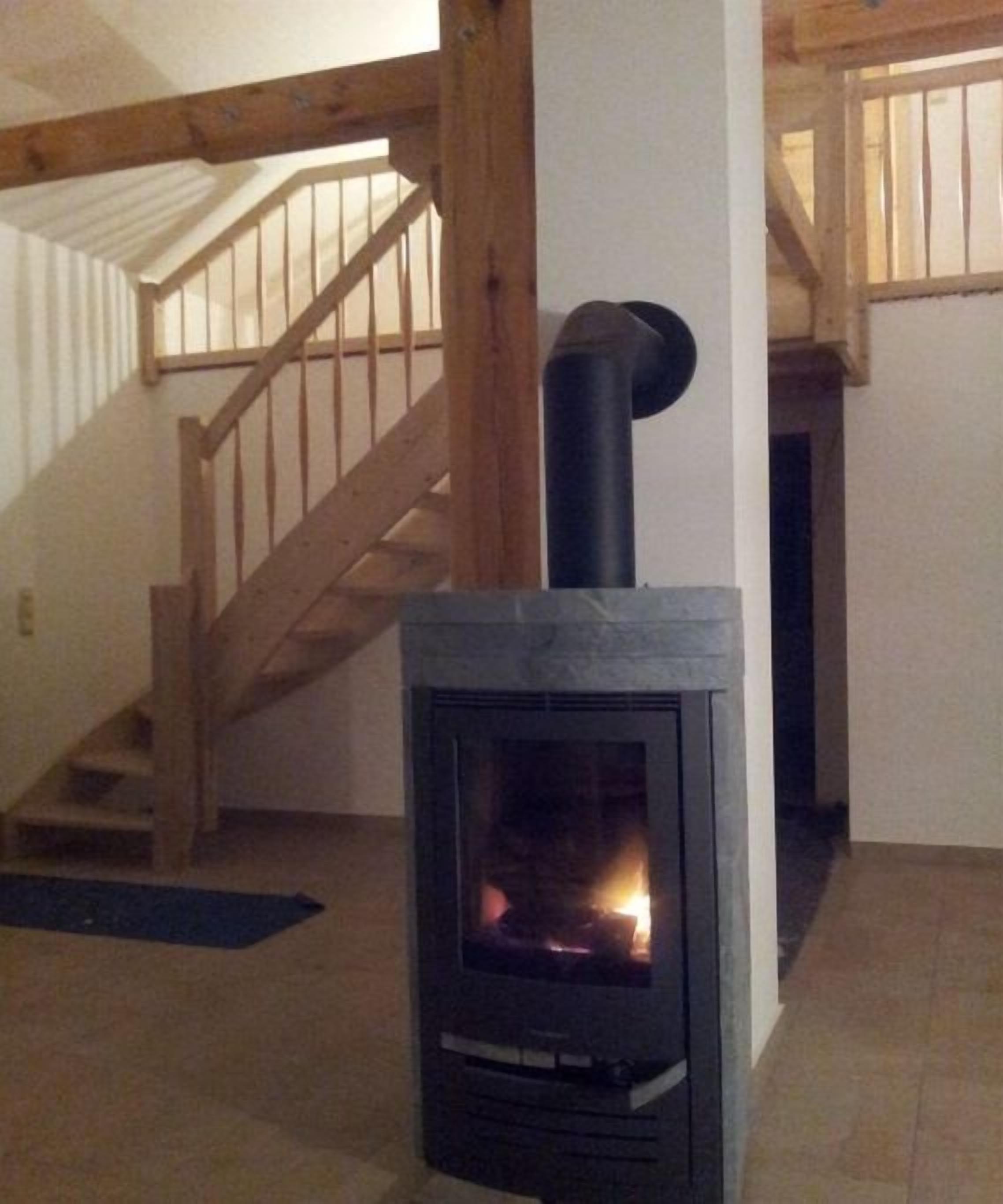 A wood-burning stove in a room with a wooden staircase and a white wall.