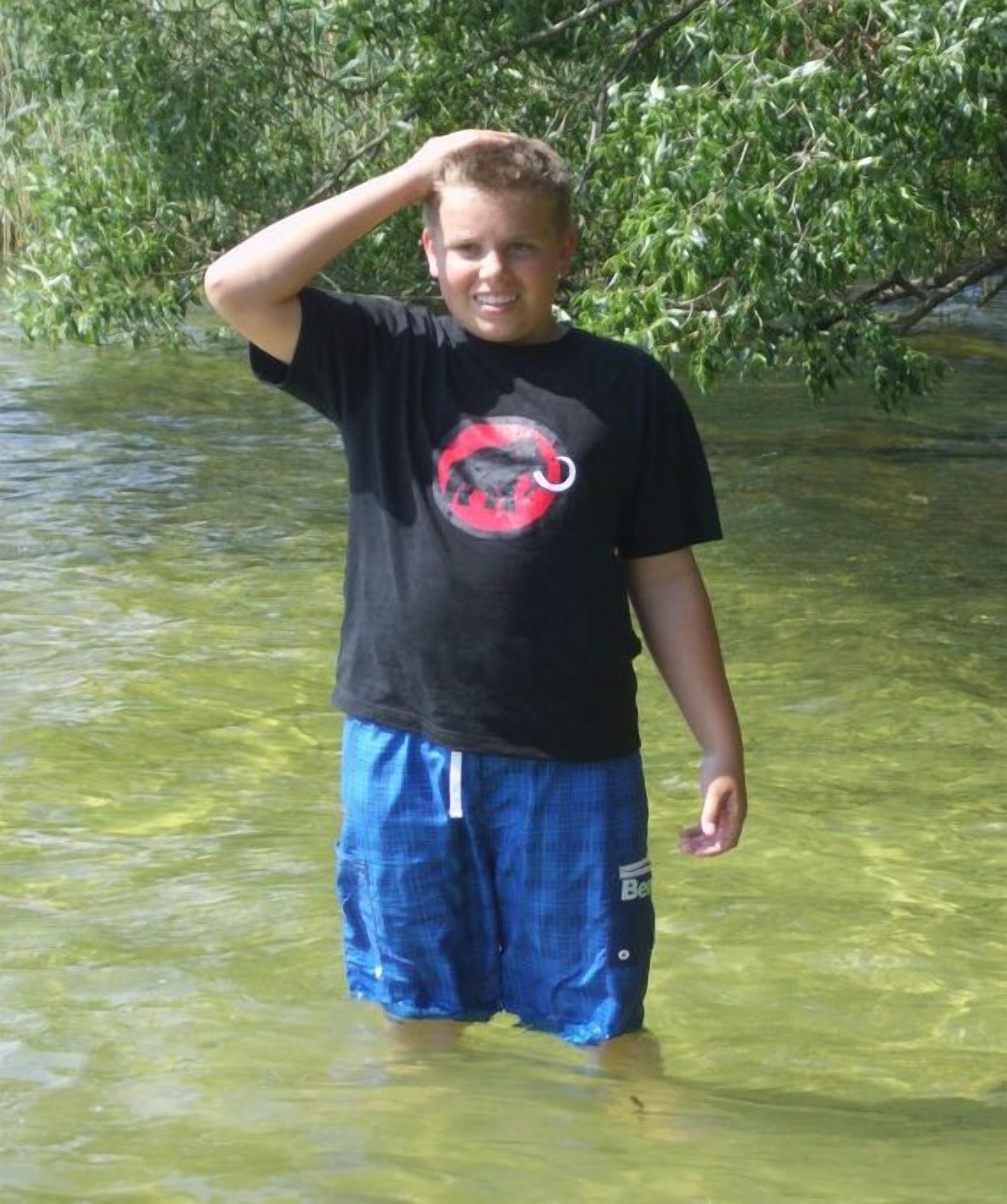Iemand staat in ondiep water, gekleed in een zwart t-shirt met een rood-witte afbeelding en blauwe korte broek.