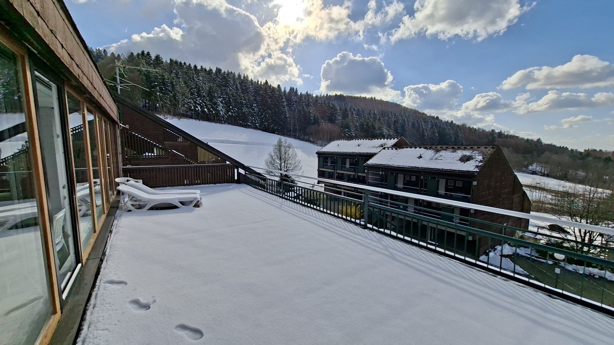 Ein mit Schnee bedeckter Balkon mit Liegestühlen, ein Holzhaus und Blick auf einen bewaldeten Hügel.