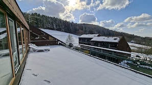 Un balcón cubierto de nieve con sillones de descanso, un edificio de madera y vistas a una colina boscosa.