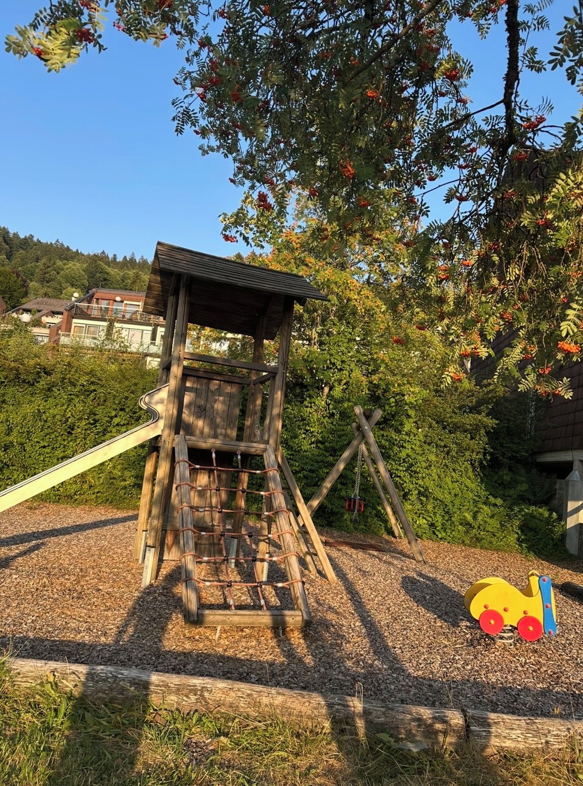 A wooden play structure with a slide and climbing net, surrounded by trees and a clear sky.