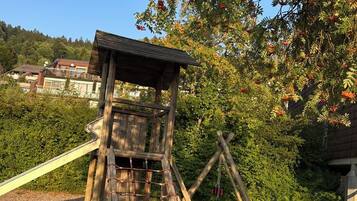 A wooden play structure with a slide and climbing net, surrounded by trees and a clear sky.