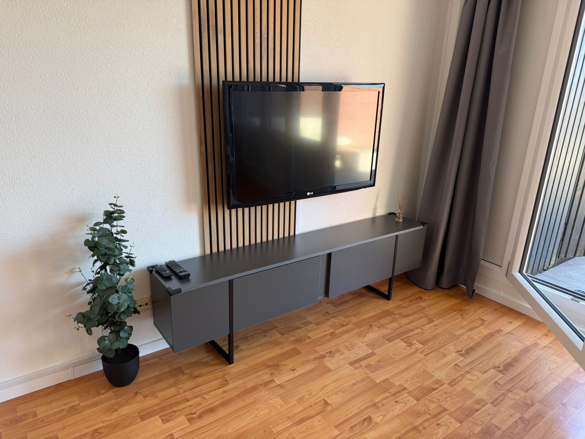Ein modernes Wohnzimmer mit einem an der Wand montierten Flachbildfernseher, einem grauen Konsolentisch, einer Topfpflanze und Holzböden.