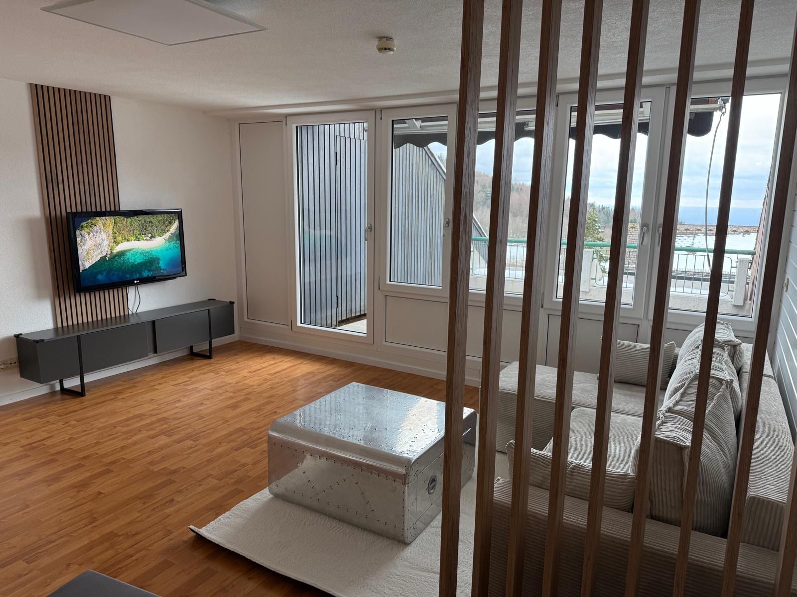 Een moderne woonkamer met een flatscreen televisie, houten vloeren, een bank en een groot raam met uitzicht op zee.