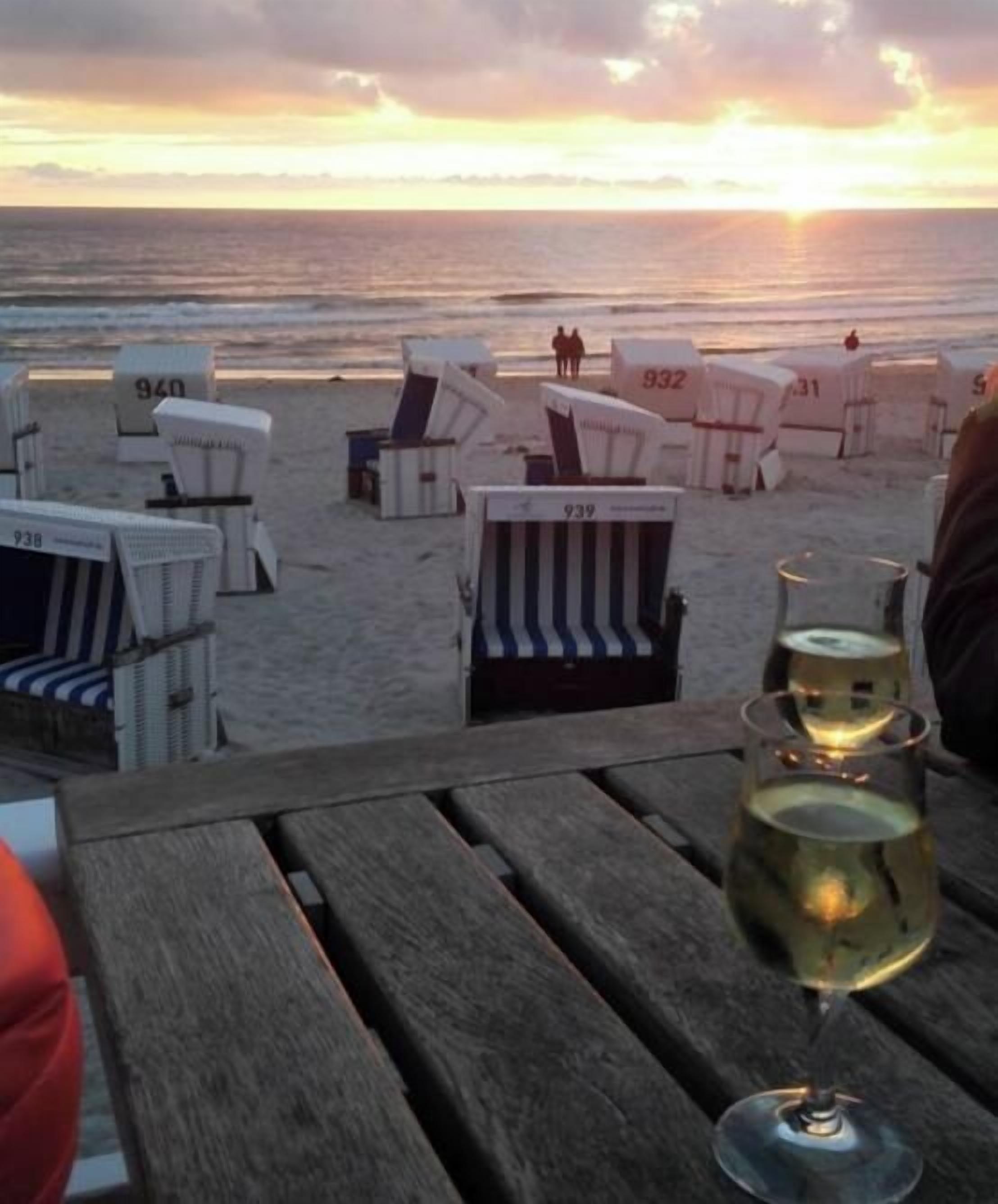 A beachside view with beach chairs numbered 939, 932, and 931, and a wooden table with a glass of white wine.