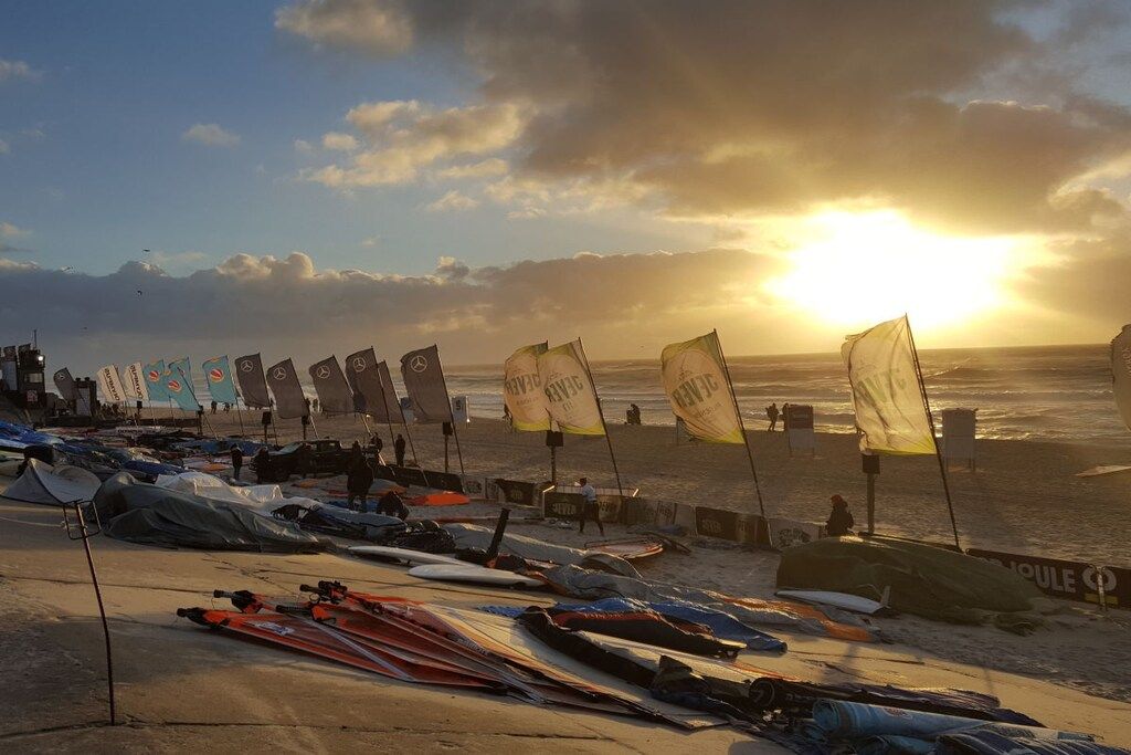 Una platja amb nombroses embarcacions de vela i banderes, sota un cel clar i amb el sol ponent sobre l'oceà.