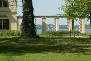 Un jardín con un pérgola, una masa de agua y un edificio con balcón.