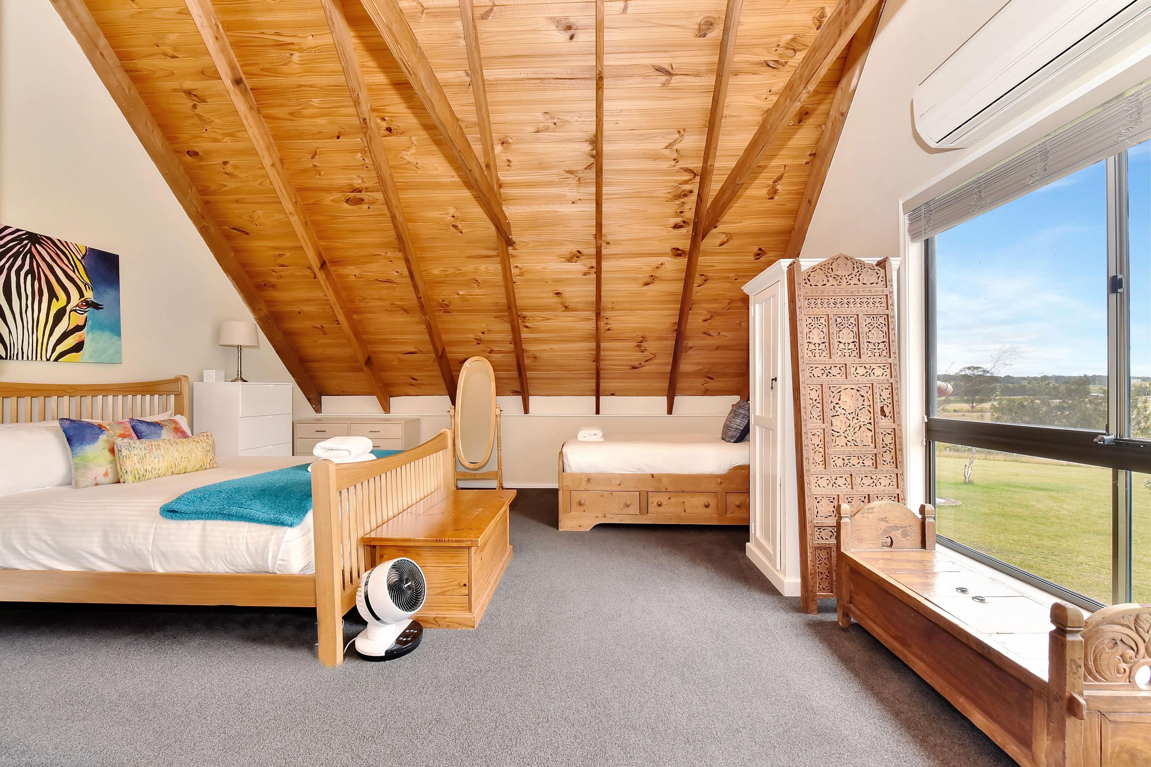 A bedroom with a sloped ceiling, a bed, a desk, a chair, and a large window with a view.