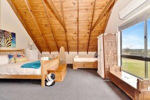 A bedroom with a sloped ceiling, a bed, a desk, a chair, and a large window with a view.