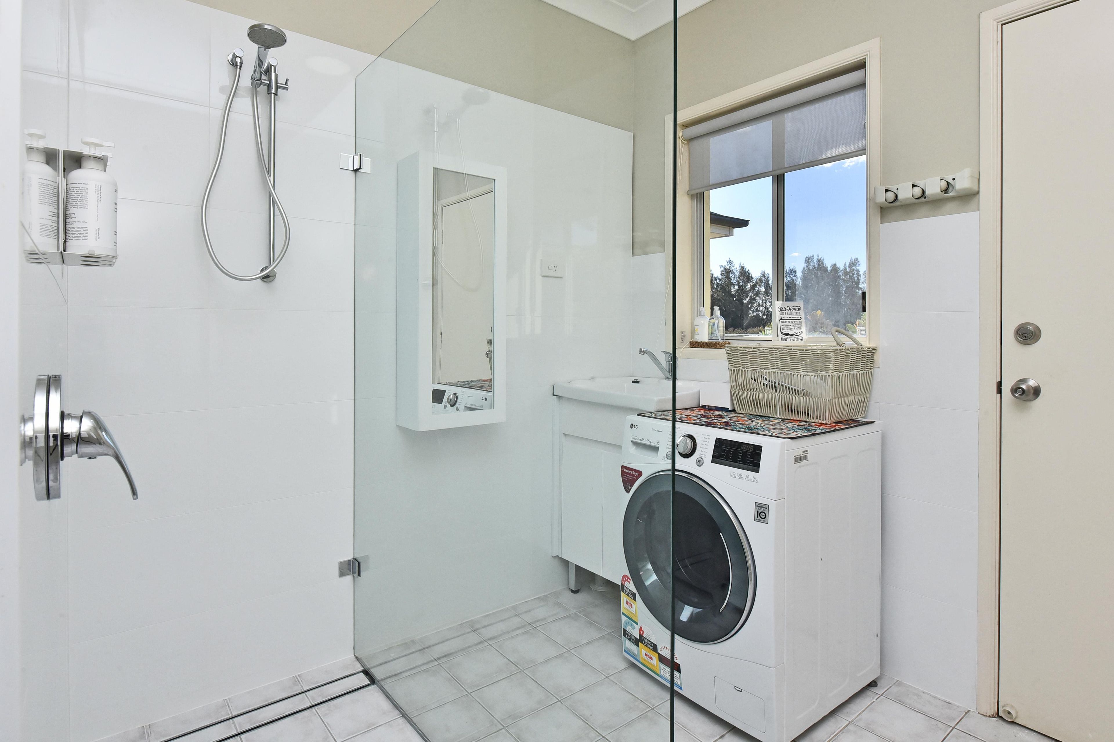 A modern bathroom with a glass shower enclosure, a built-in washing machine, and a window providing a view.
