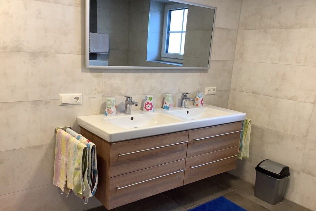 A bathroom with a double sink vanity, wooden drawers, a mirror above the sink, and a trash bin.