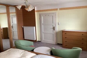 Ein Schlafzimmer mit einem Bett, zwei grünen Stühlen, einer Holzkommode, einem Heizkörper und einer weißen Tür.
