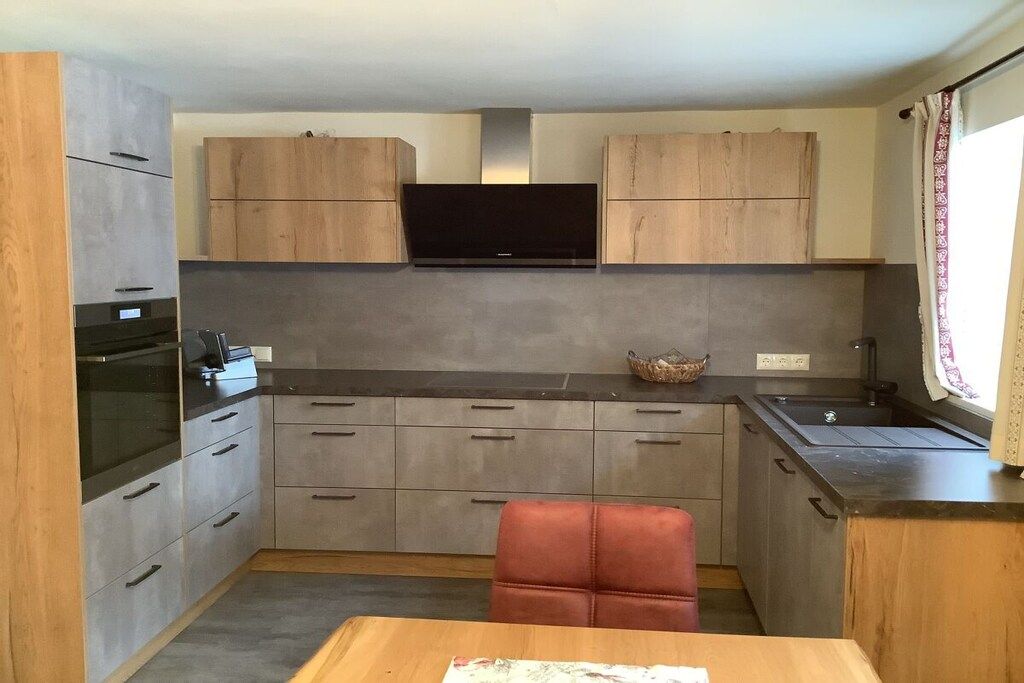 A compact kitchen with grey cabinets, a black range hood, a built-in TV, and a small dining area with a red chair.
