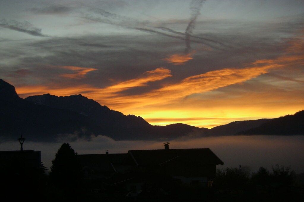 Ein Berglandschaft im Abendrot mit Gebäude-Silhouetten und einem Gewässer im Vordergrund.