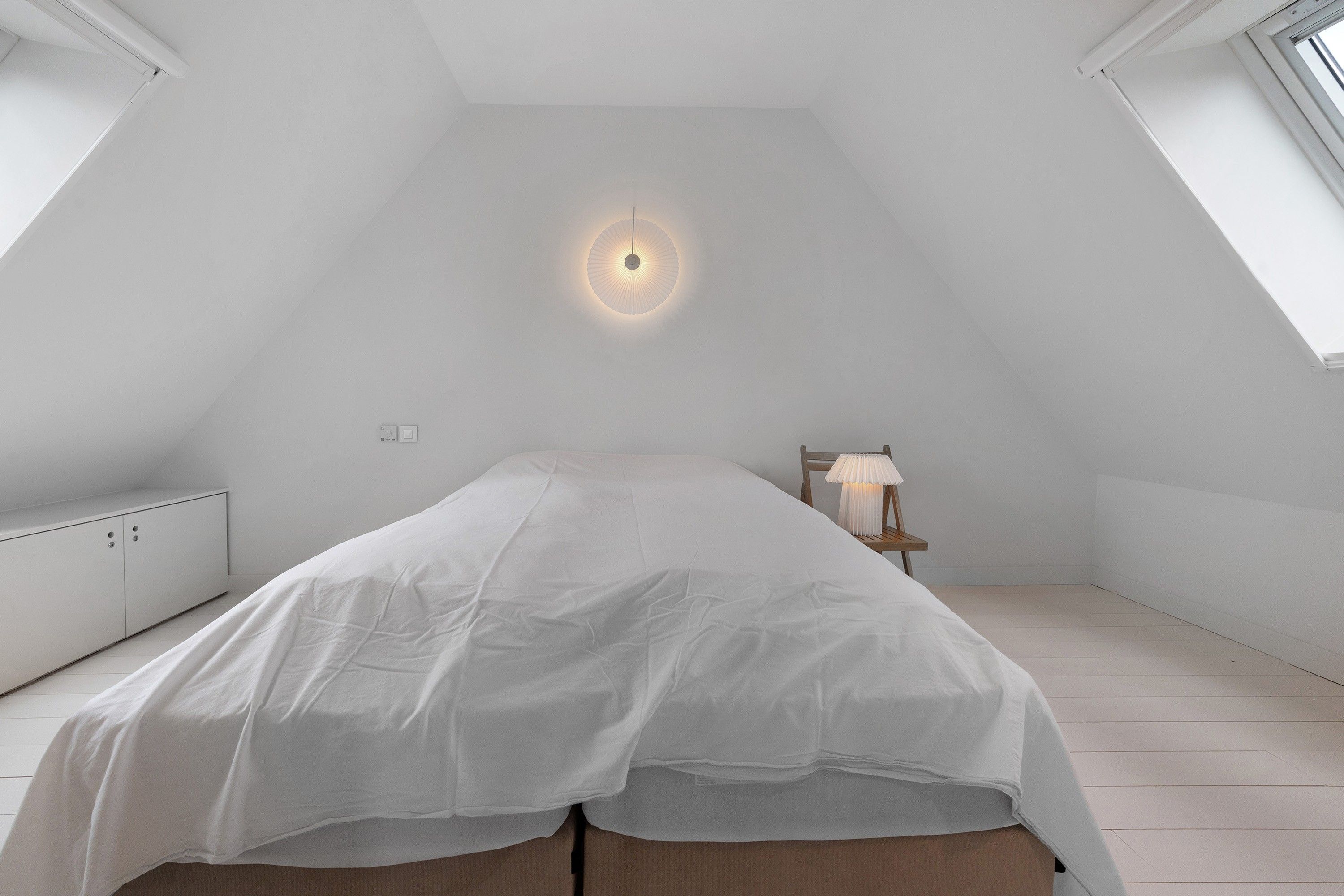 A bedroom with a sloped ceiling, a bed with a white bedspread, a nightstand with a lamp, and a window.