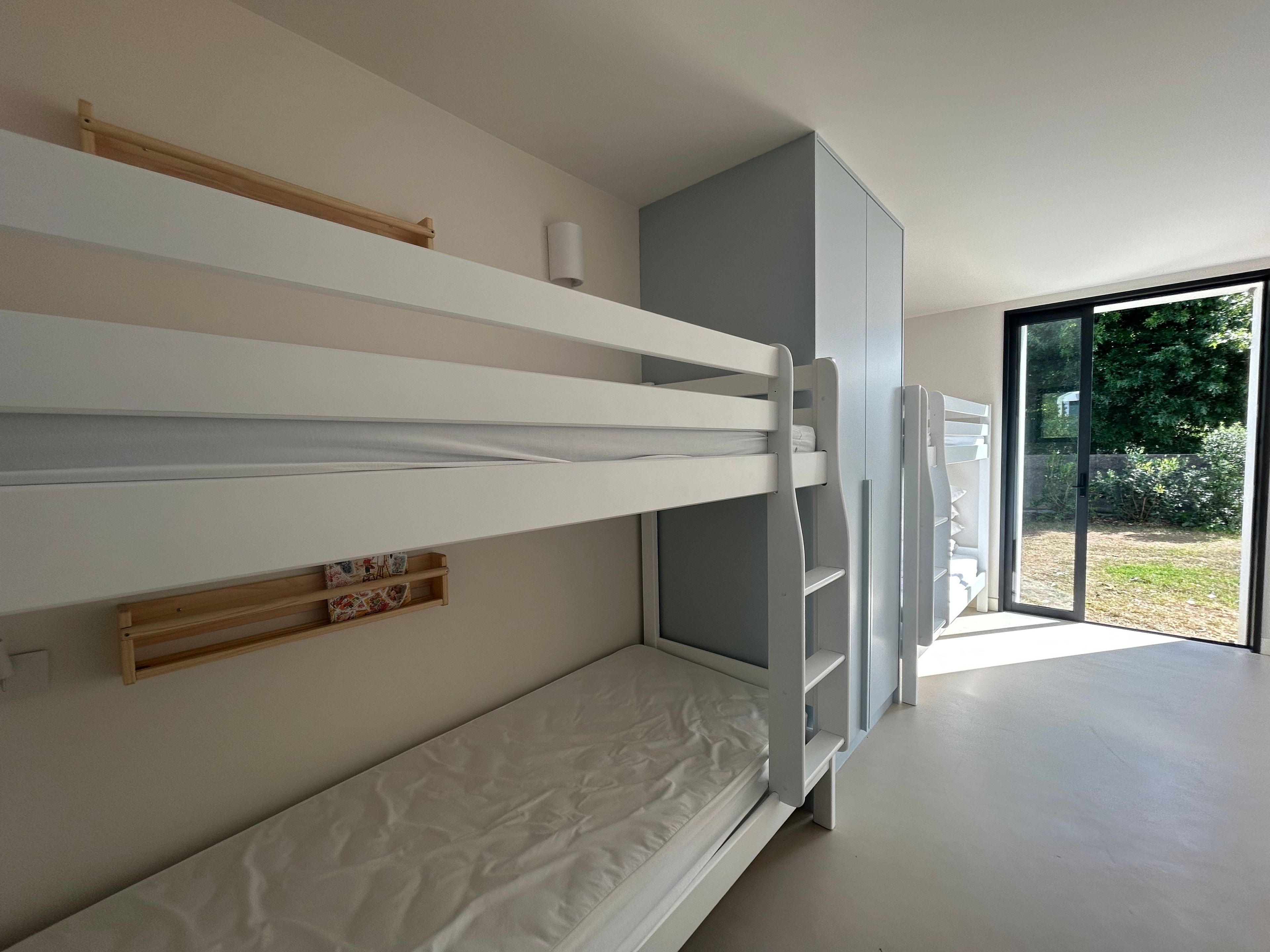 Una cama individual superpuesta con un estante de madera y una mesita, una puerta corredera a un balcón y vistas de vegetación.