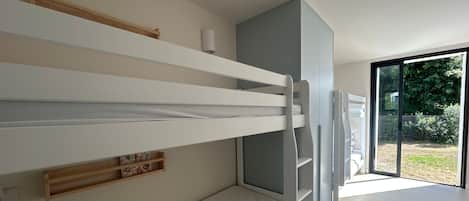 Un lit superposé avec une étagère en bois et une petite table, une porte coulissante donnant sur un balcon et une vue sur la verdure environnante.