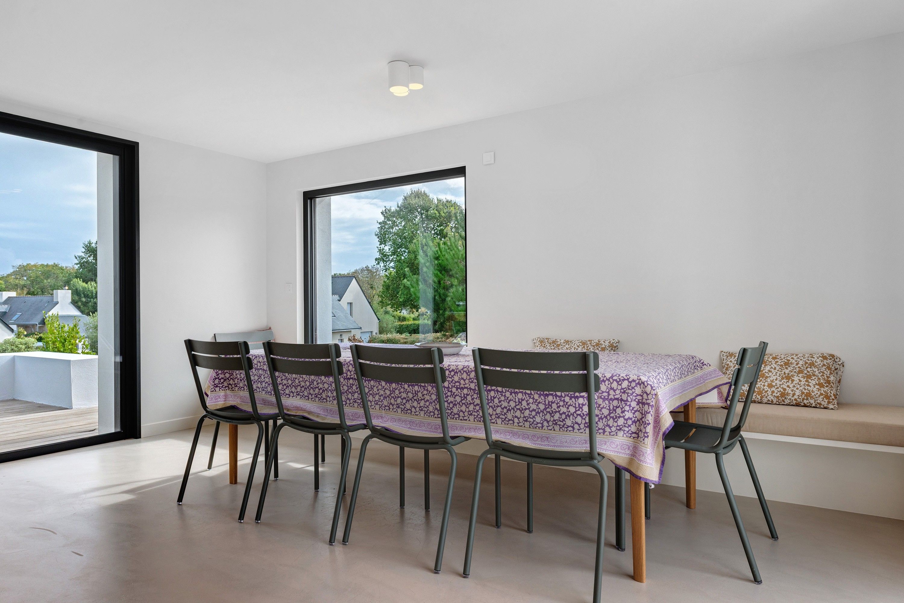 A dining area with a table and chairs, a large window, and a view of greenery outside.