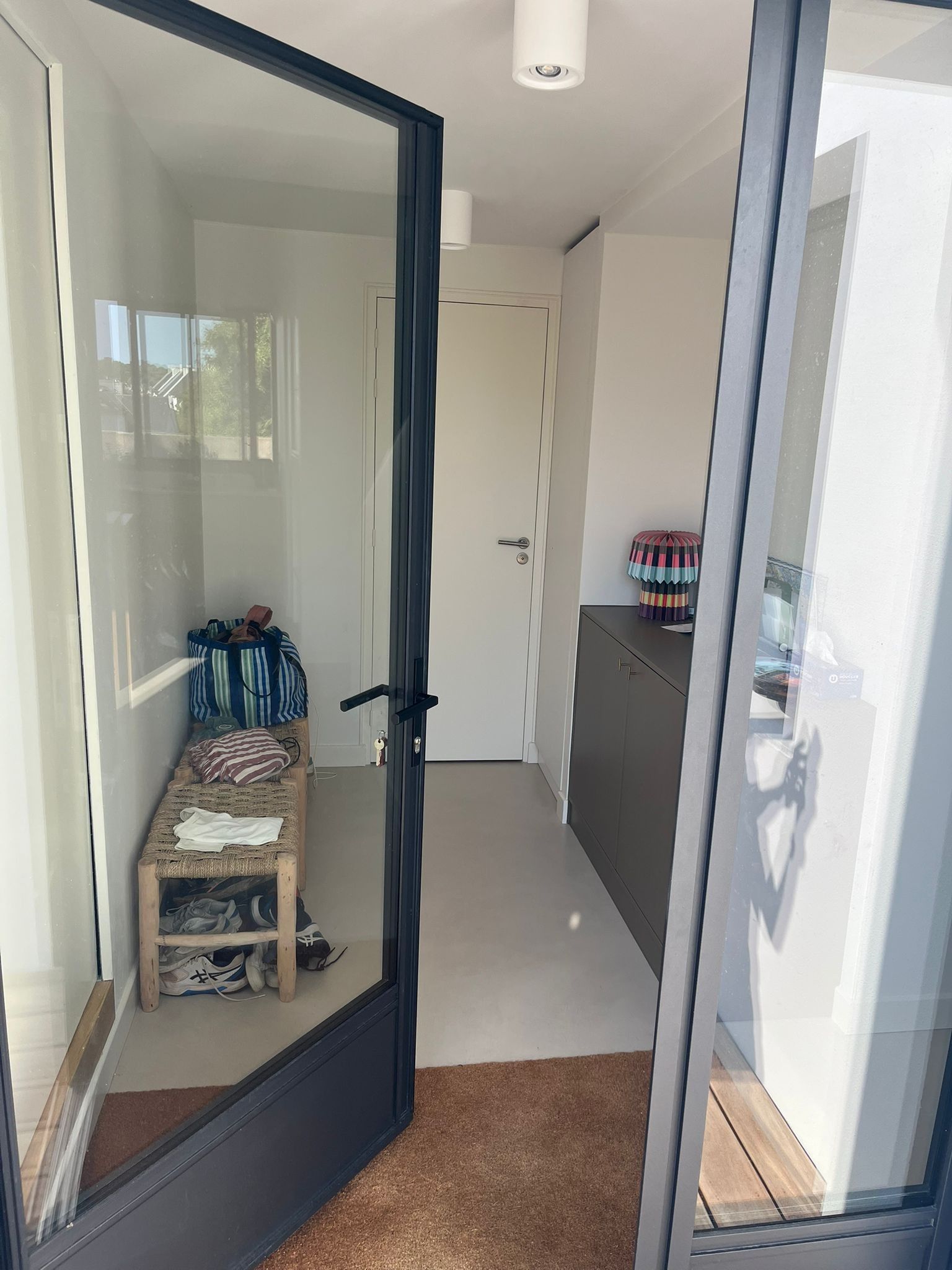 Una habitación con puerta de vidrio que da a un balcón, un pequeño banco con cojín, un par de zapatos y un cojín de colores sobre un mueble.