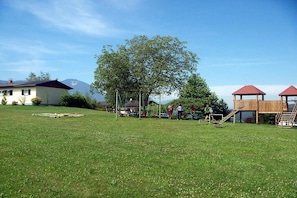 Ein grünes Areal im Freien mit einem Spielplatz, darunter eine Rutsche und Schaukeln, in der Nähe eines Gebäudes mit rotem Dach.