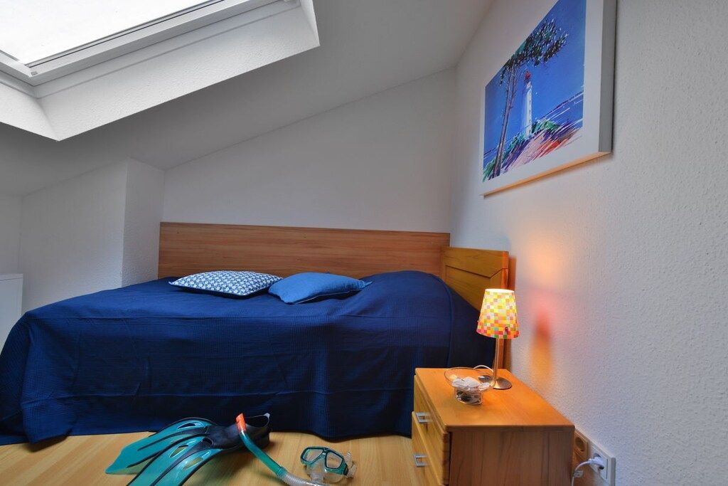 Quarto com claraboia, cama com roupa de cama azul, mesa de cabeceira com abajur e pintura de um farol.