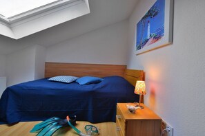 A bedroom with a skylight, a bed with blue bedding, a bedside table with a lamp, and a painting of a lighthouse. - Holiday apartment in Juliusruh with parking (Juliusruh)