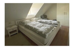 A bedroom with a sloped ceiling, a bed with white bedding, a nightstand, and a wardrobe. - Holiday apartment with 2 bedrooms (Utersum auf Föhr)