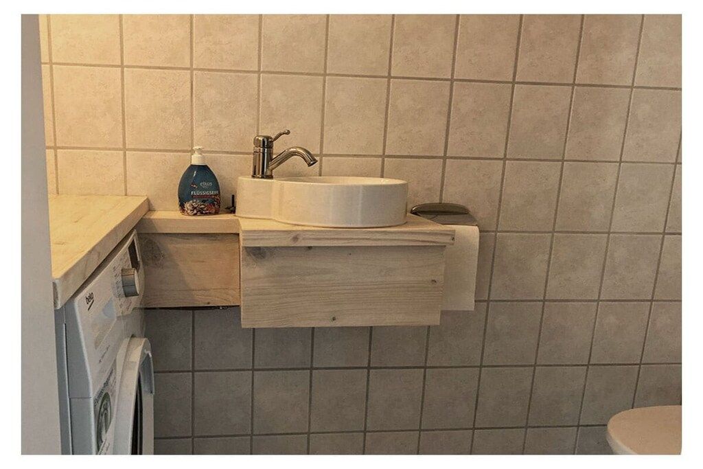 A bathroom with a white sink, a soap dispenser, and a wooden shelf.