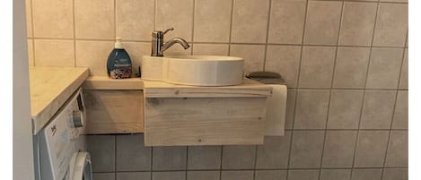 Un baño con un lavamanos blanco, un dispensador de jabón y un estante de madera.