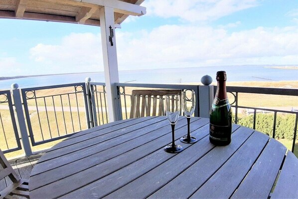 Une table en bois, une bouteille de champagne et deux verres sont posés sur un balcon donnant sur une étendue d’eau.