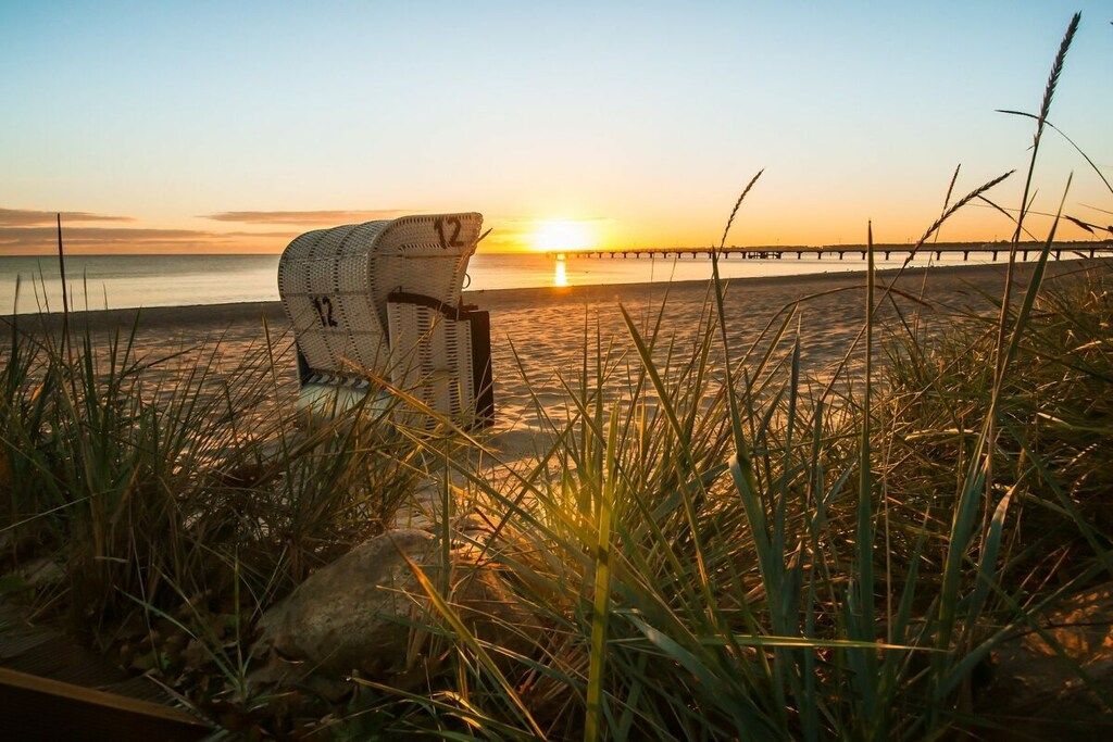 A beach chair on the sand with a view of the sunset and a pier in the background.