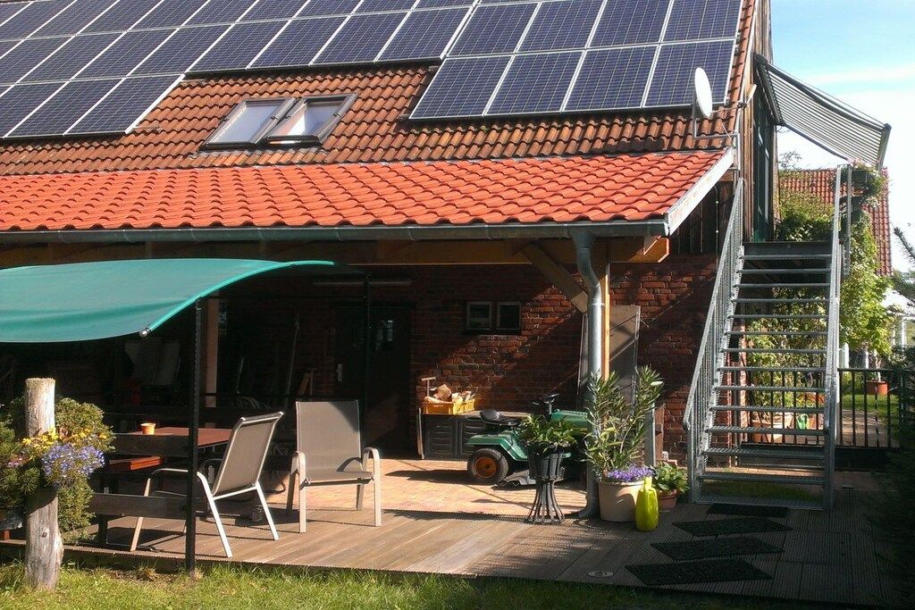 Une maison avec un toit recouvert de tuiles rouges, des panneaux solaires, un patio couvert avec du mobilier et une façade en briques.
