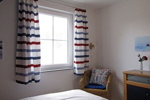 Ein Schlafzimmer mit einem Bett, einem Korbstuhl, einem Nachttisch aus Holz und einem Fenster mit Vorhängen.