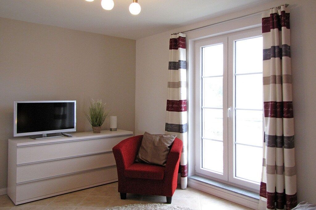Un salon avec un téléviseur à écran plat posé sur un meuble blanc, un fauteuil rouge avec un coussin, une plante en pot et une fenêtre ornée de rideaux à rayures.