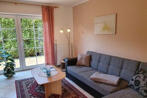 A living room with a sofa, a table, a radiator, and a window with curtains. - at Vanessa's House || Zingst (Zingst)