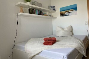 A bedroom with a bed, a white wall, a shelf with books and a plant, and a painting of a beach scene. - Holiday apartment with 2 bedrooms (Scharbeutz\/Haffkrug)