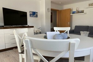 Ein modernes Wohnzimmer mit einem Flachbildfernseher, einem runden Esstisch mit Stühlen, einem grauen Sofa und einer Holztür.